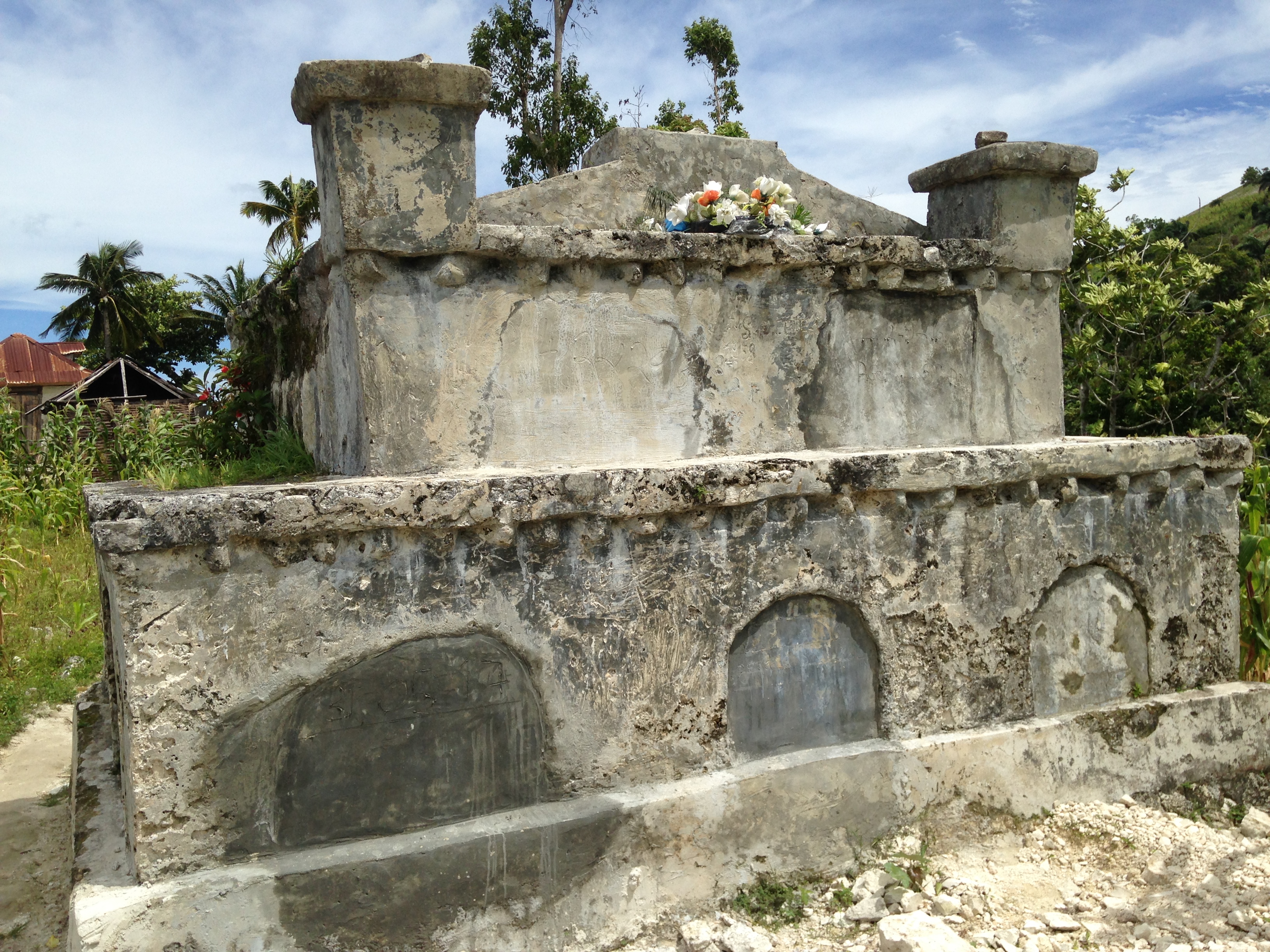 Color photograph of a large tomb for an ancestor in Delatte, Haiti, near the western coastal city of Petit-Goave. Two-tiered cement structure, roughly four feet high, adorned with plastic flowers, and surrounded by green overgrowth in a yard.