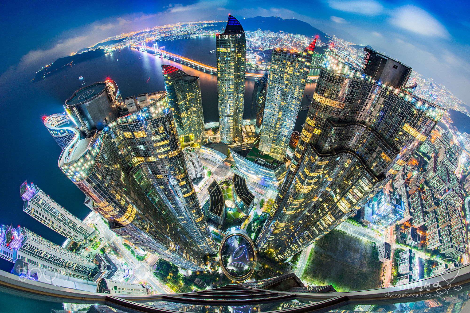 Color photograph taken at night from the eighty-eighth floor of a high rise, looking down upon other buildings and a well-lighted modern cityscape.