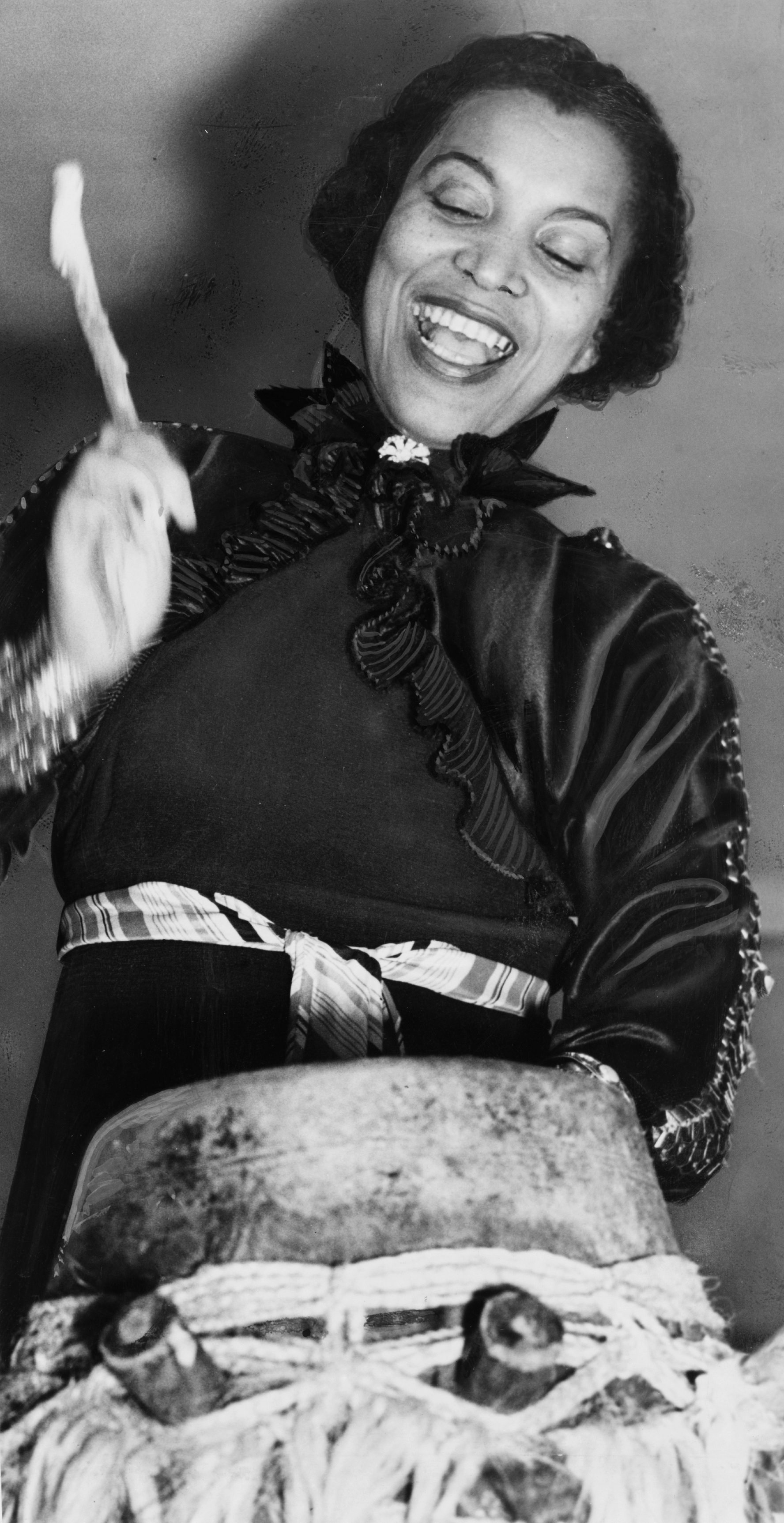 Nineteen thirty-seven black and white photograph of American author and anthropologist Zora Neale Hurston, who is here standing and beating with a stick a large Vodou drum, while she smiles.