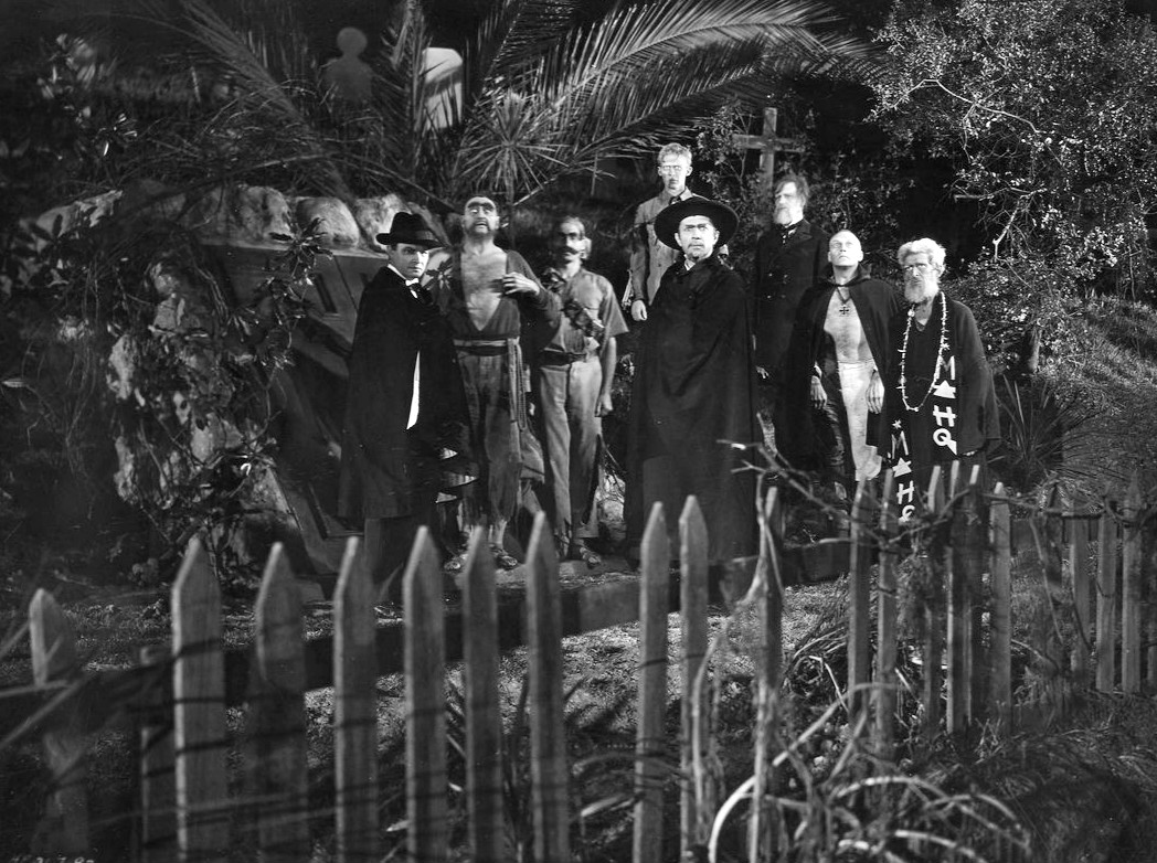 Nineteen thirty-two black and white photograph of a scene from the first ever zombie movie, White Zombie, depicting the villainous zombie master, Murder Legendre (played by Bella Lugosi), surrounded by several of his white zombies in a cemetery in Haiti, a decrepit, unpainted picked fence before them.
