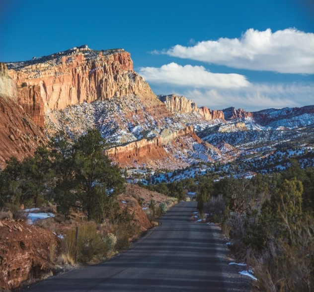 Blacktop road with small green trees and shrubs and red cliffs covered in snow, with blue sky and clouds above. 