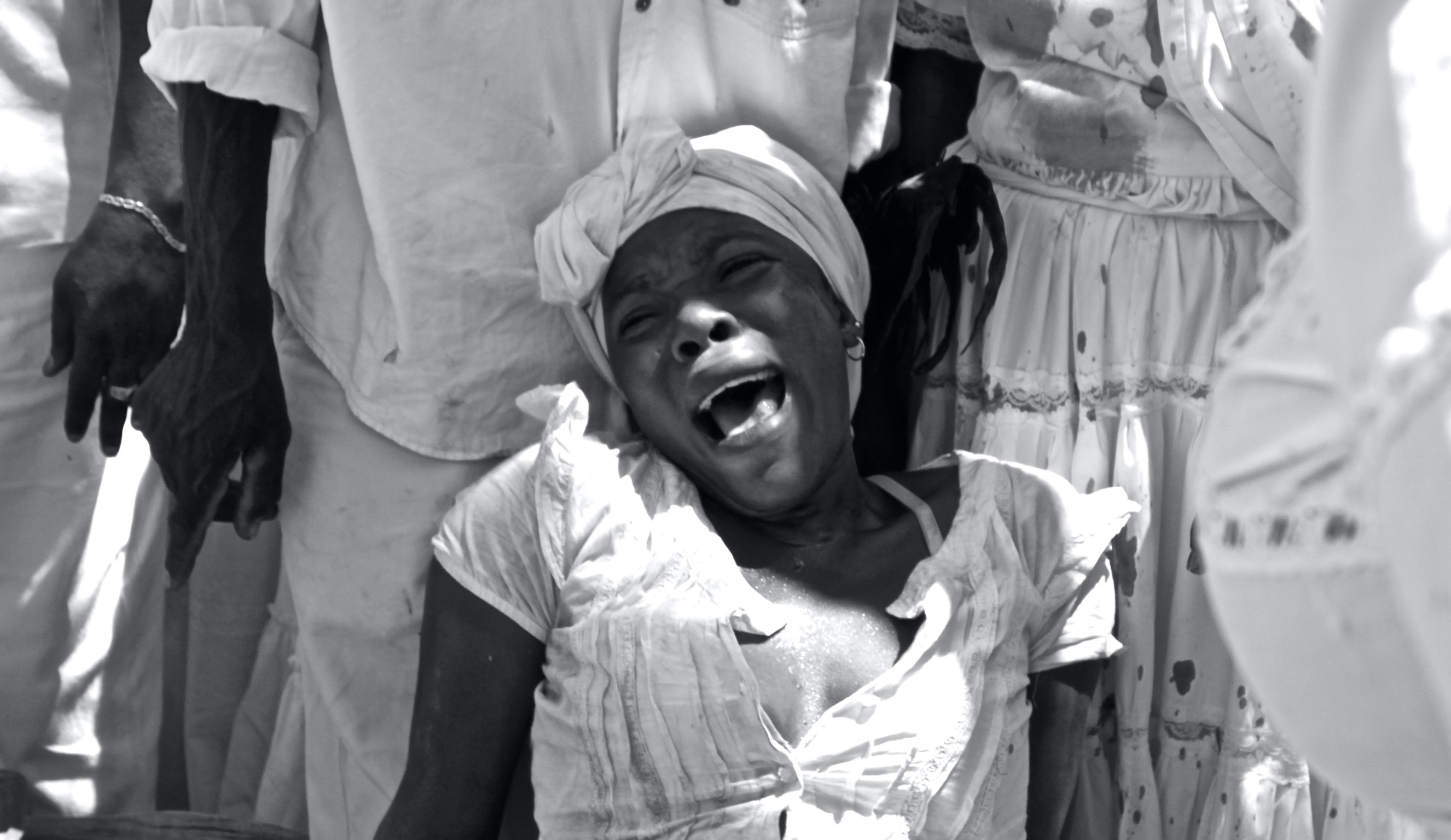 Black and white photograph of a woman, perhaps a priestess, being possessed by a spirit at a Vodou ceremony in central Haiti, by now seated on the floor behind other devotees and deeply engaged with divinity.