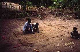 Color photo of children in a yard playing a soccer match with bottle caps and a round seed, the ball, on a traced out field in the dirt yard.